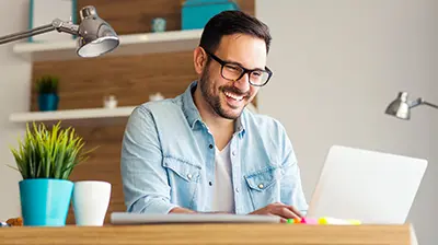 A man in casual attire sitting at his desk while laughing and using his laptop 