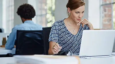 A woman sitting at a desk and using a laptop while taking notes. A man is sitting at a desk in the background  while working. 