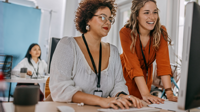 Three women in a office at their desks. One woman is sitting at her desk using her computer while another woman in leaning on the desk and looking at the same computer 