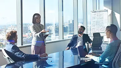 Group of men and women in business attire in a conference room in an office, the woman is standing and the men are sitting around a large table looking up at her.
