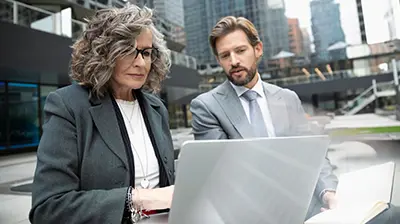 Woman and man colleagues  dressed in business attire sitting outside office buildings conversing and looking at a laptop.