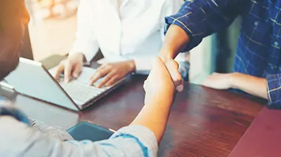 A handshake happening across a table. There is a person on a laptop in the background