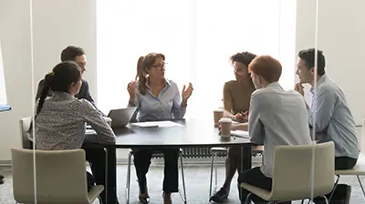 Six people sitting around a table having a meeting. A woman in the middle is the main speaker while the others are listening.